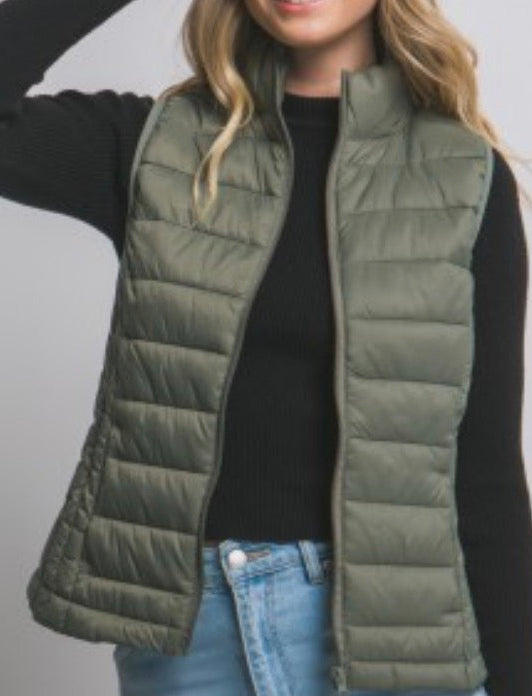 Woman wearing a green puffer vest, black long-sleeve top, and light blue jeans on a gray background